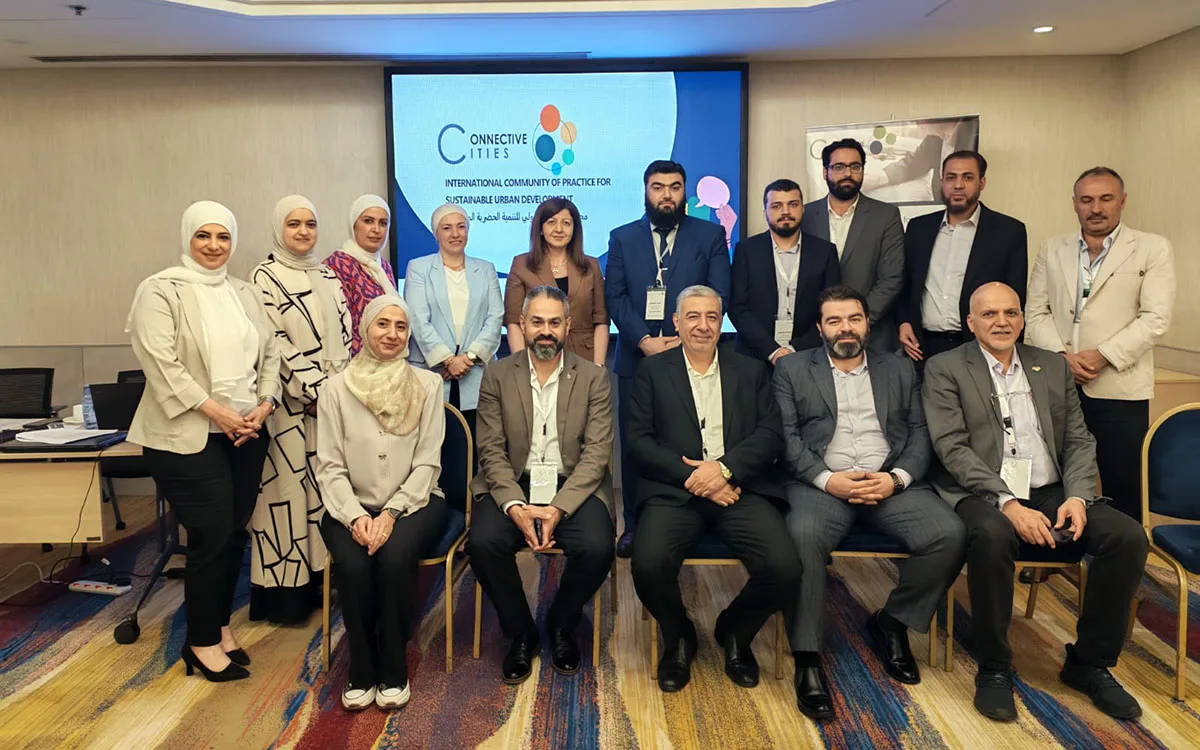 The participants, some standing and some seated in the conference room, with a screen displaying the Connective Cities logo in the background.