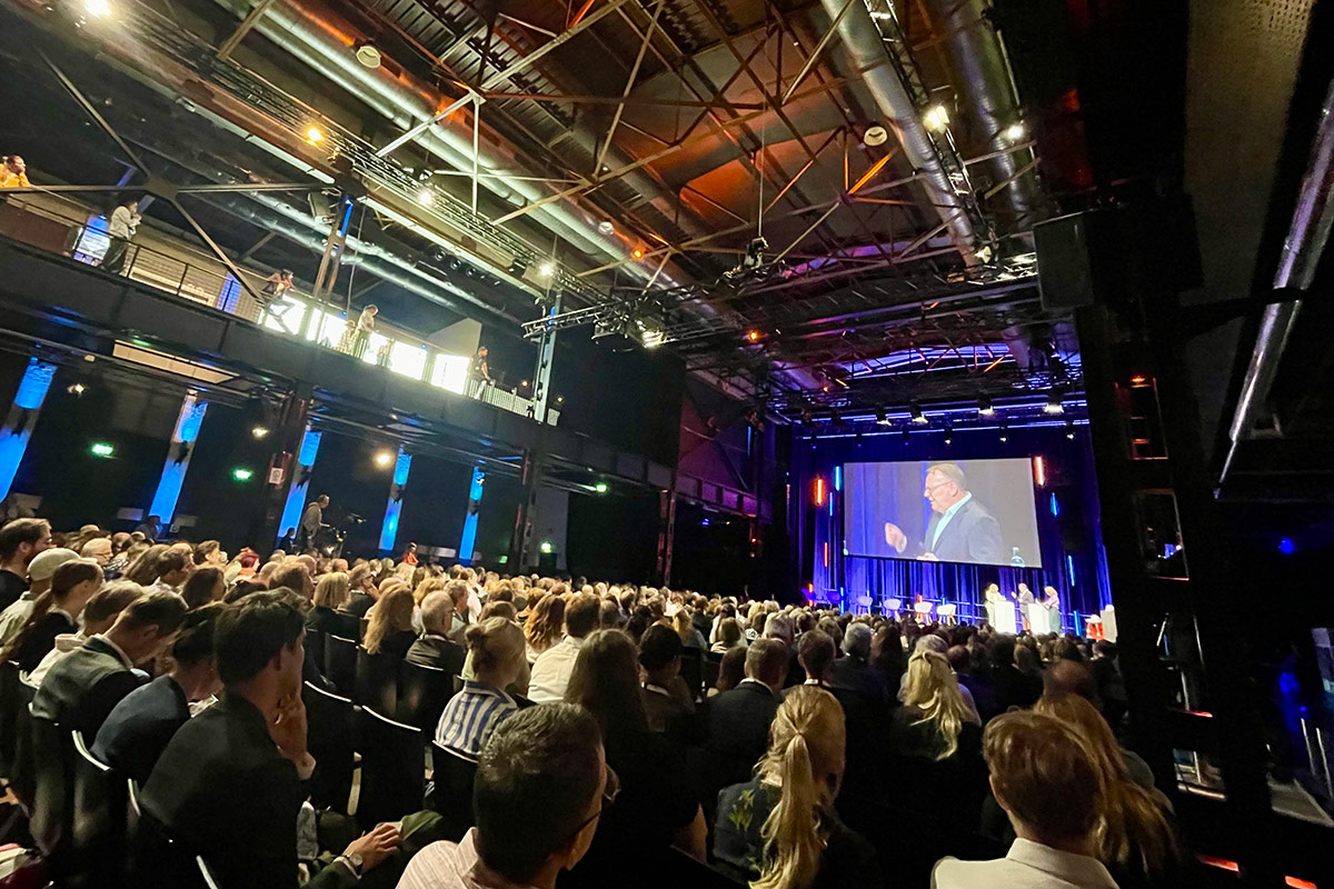 An industrial-style conference room bathed in violet-blue light. At the back of the stage is a large screen showing a picture of Torsten Burmester; in the foreground, the room is packed with participants.