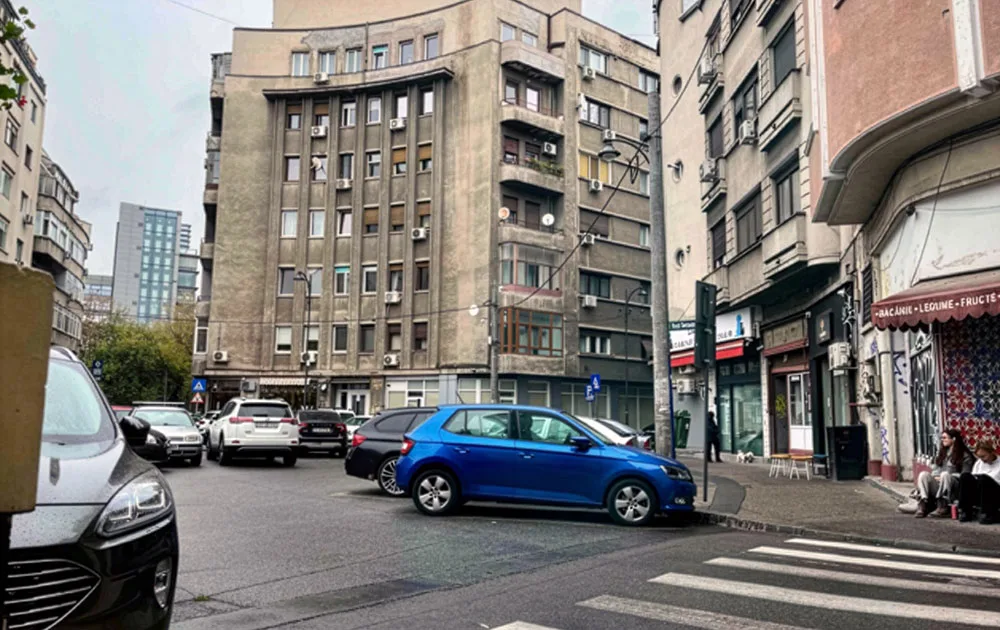 An old high-rise building in Bucharest. In front of it, a paved square filled with parked cars. 
