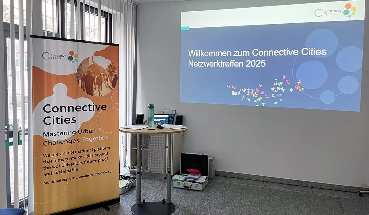 Links im Bild ist ein Banner von Connective Cities und mittig ein Infoscreen mit einem Schriftzug "Willkommen zum Connective Cities Netzwerktreffen 2025".