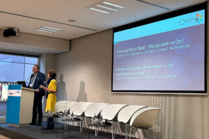 Thomas Otto, Bürgermeister der Gemeinde Saterland, spricht am Rednerpult. Daneben steht Sibylle Loyeau, Projektleiterin von Connective Cities. Auf der Leinwand: der deutsch-ukrainische Titel „Praxis aus Deutschland – Was passiert vor Ort?“.