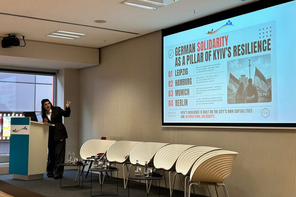 Dinara Habibulaieva, Stadträtin in Kyjiw, spricht am Pult und zeigt auf die Leinwand. Die Folie „German Solidarity as a Pillar of Kyiv’s Resilience“ nennt Hilfen aus Leipzig, Hamburg, München und Berlin.