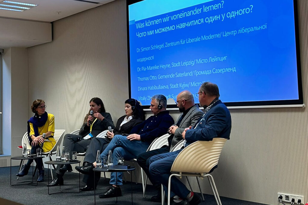 Podiumsdiskussion „Was können wir voneinander lernen?“: Sechs Personen sitzen mit Mikrofonen auf der Bühne, eine Frau in Gelb links. Auf der Leinwand stehen Titel und Namen der Beteiligten auf Deutsch und Ukrainisch