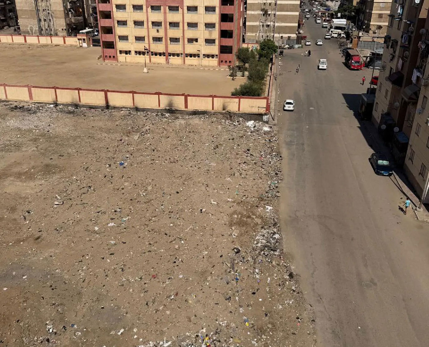 Blick von oben auf eine große, sandige Brachfläche mit verstreutem Müll neben einer Straße. Dahinter liegen eine ummauerte Freifläche mit einem mehrstöckigen Gebäude sowie Wohnhäuser.